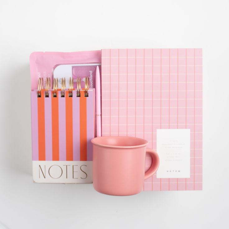 a pink coffee cup sitting on top of a desk next to a notepad and pen