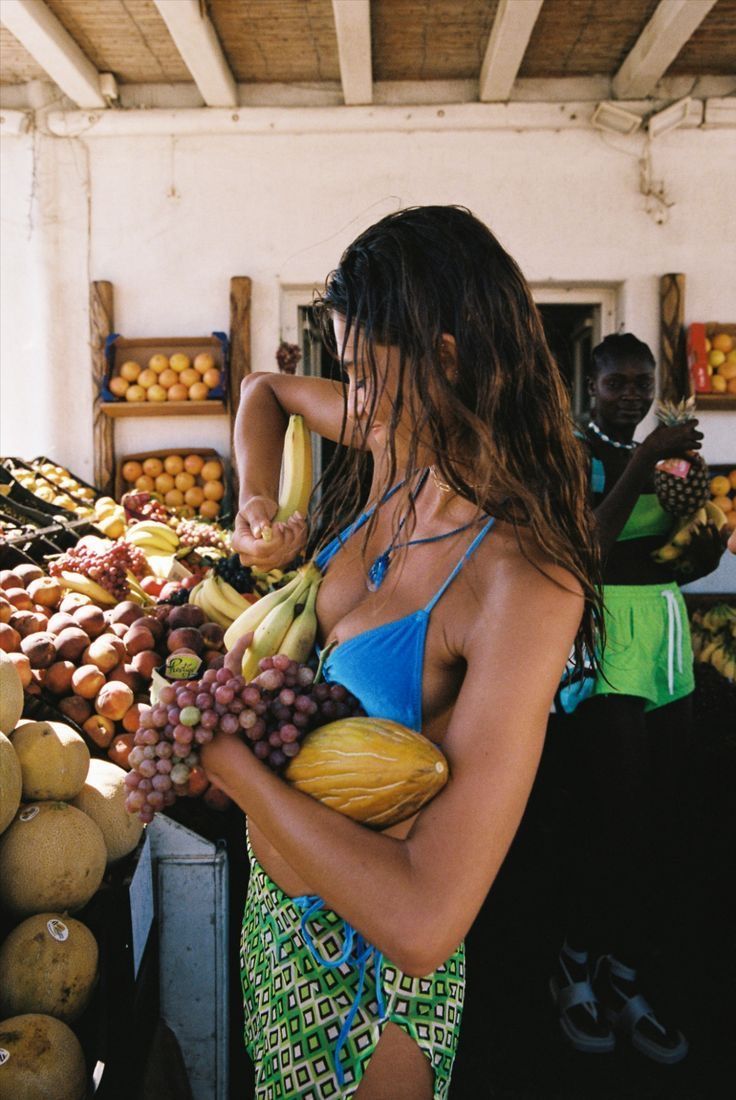 a woman standing in front of a fruit stand holding bananas and other fruits on display