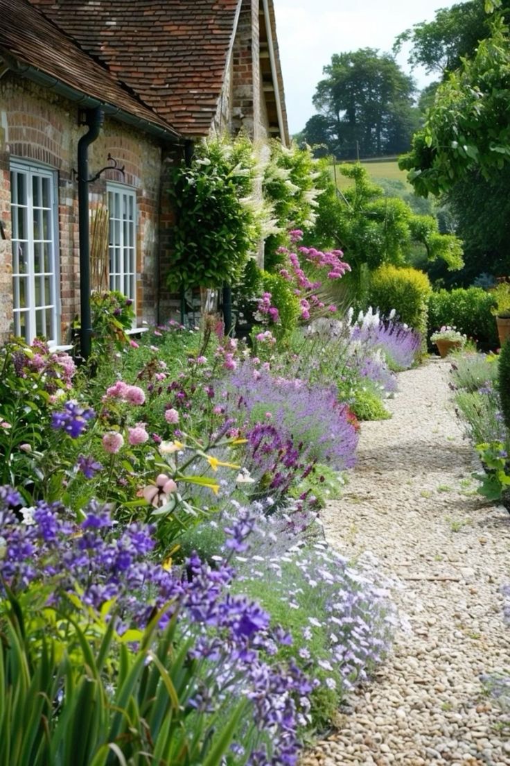 a garden with lots of flowers next to a brick building and stone walkway leading up to the front door