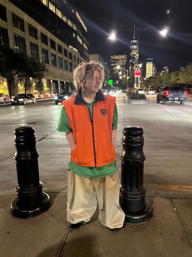 a young man standing on the side of a street next to two black poles with lights in the background