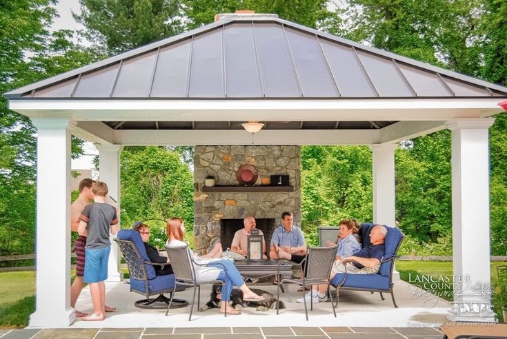 a group of people sitting around a table under a gazebo on a patio with an outdoor fireplace