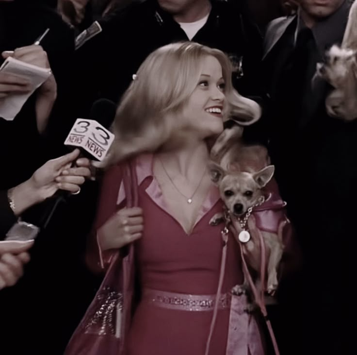 a woman in a red dress holding a dog and talking to the camera while surrounded by people