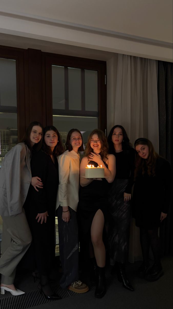 a group of women standing next to each other in front of a window holding a cake