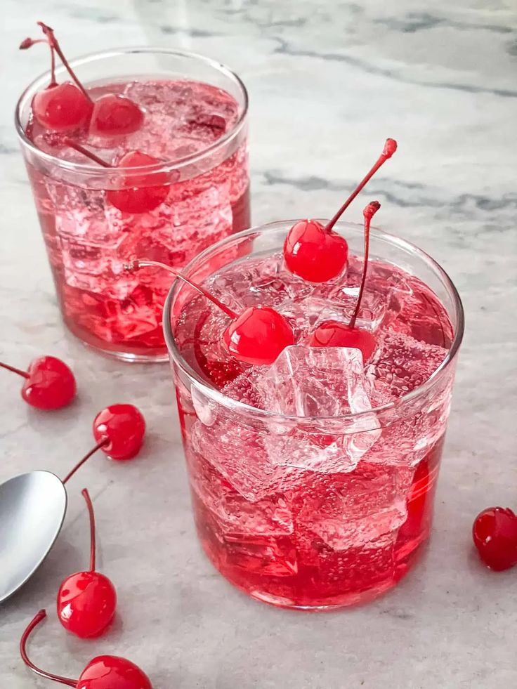 two glasses filled with ice and cherries on top of a marble table next to spoons