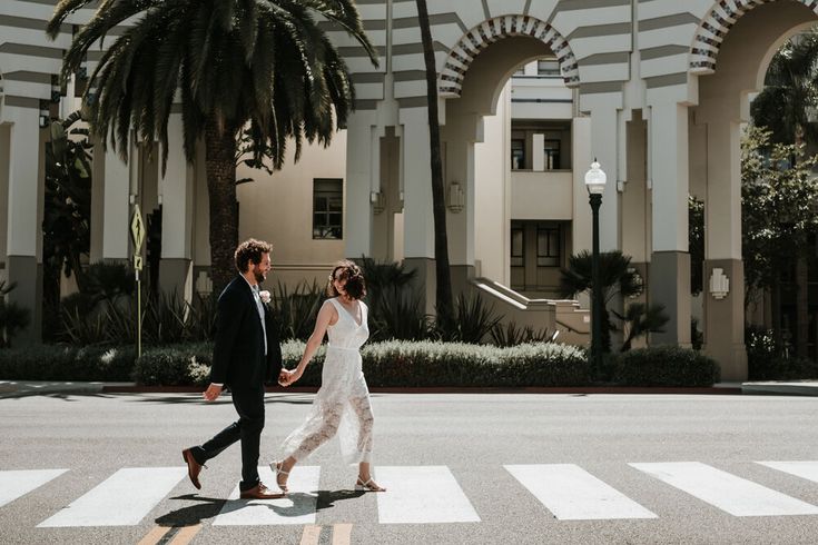 a bride and groom crossing the street in front of an elegant building with palm trees