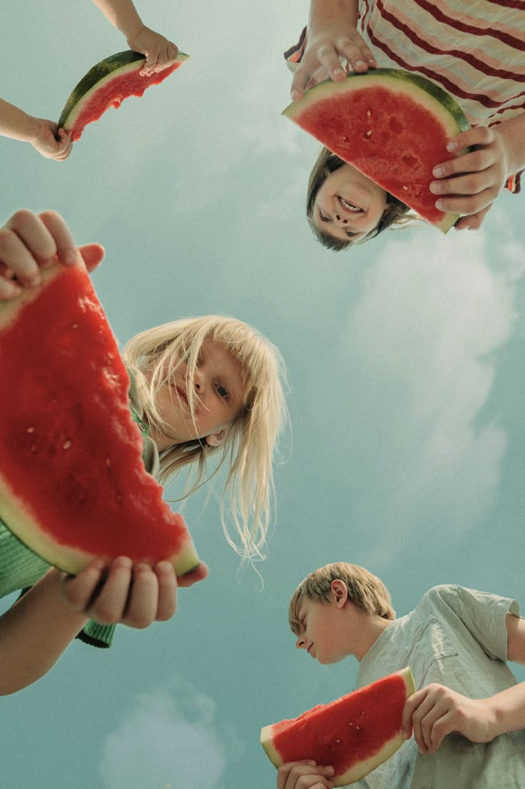 four children holding slices of watermelon in front of their faces and looking up at the sky