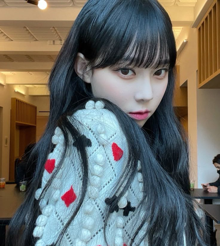 a woman with long black hair and red hearts on it's sweater is looking at the camera