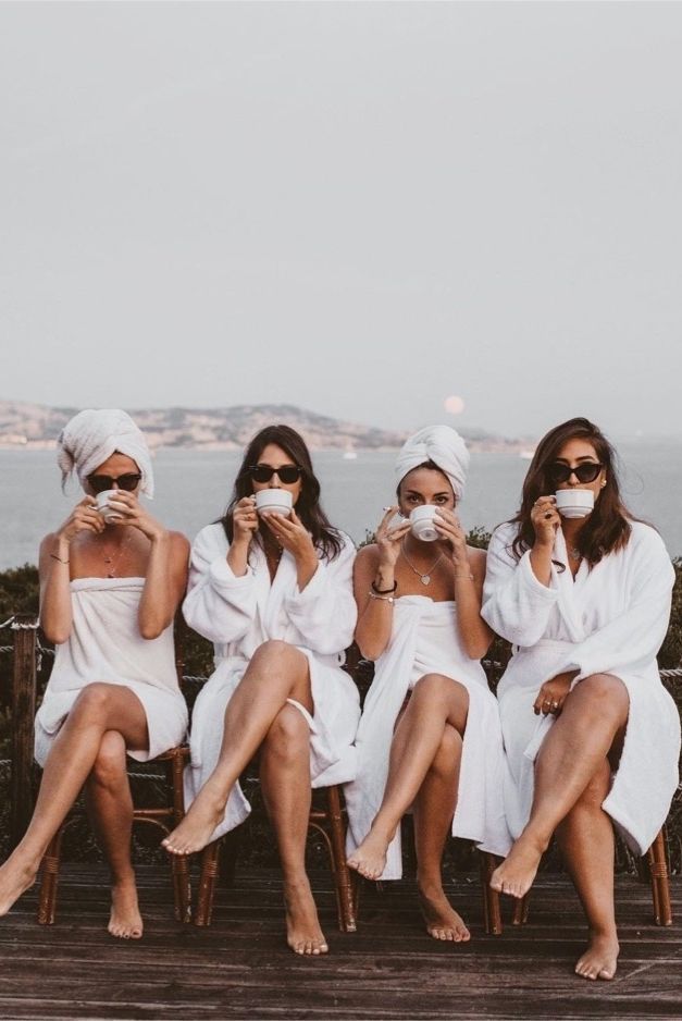 three women in robes sitting on a bench with their faces covered by towels and drinking coffee