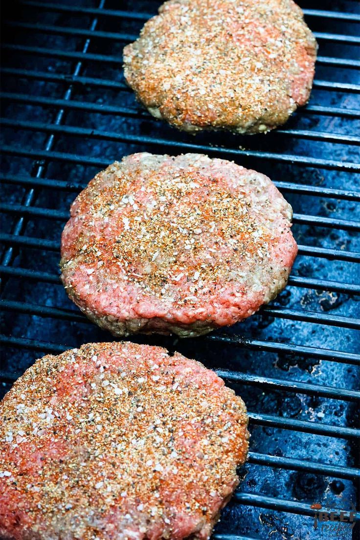 three hamburger patties cooking on a grill with seasoning sprinkled on top