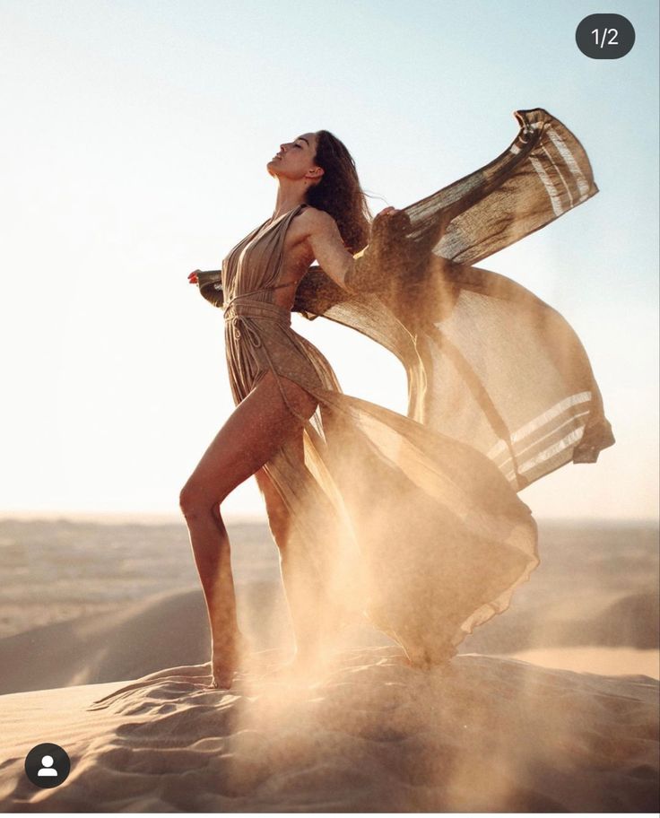 a woman standing in the sand with her arms around her body and holding a scarf over her head