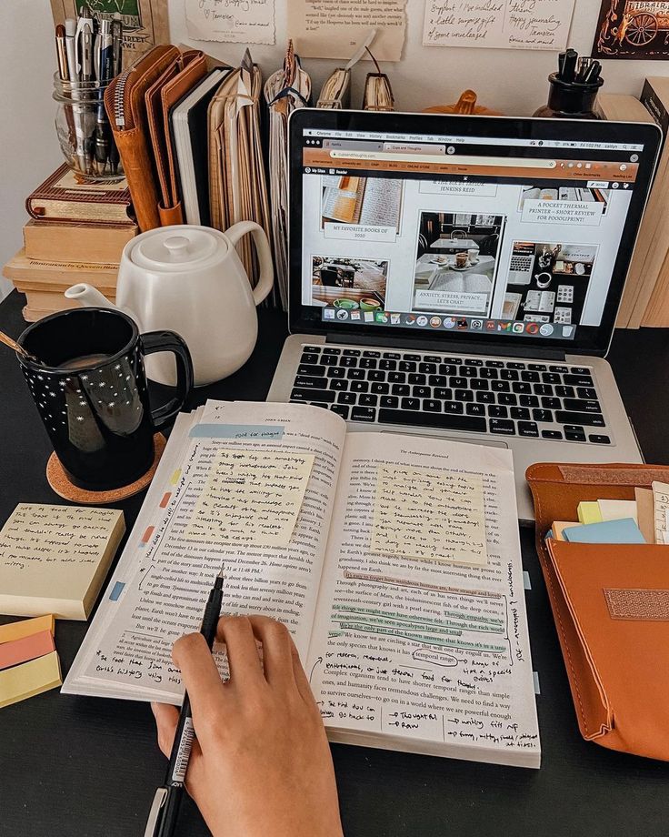 a person is writing on an open book in front of a laptop and coffee mug