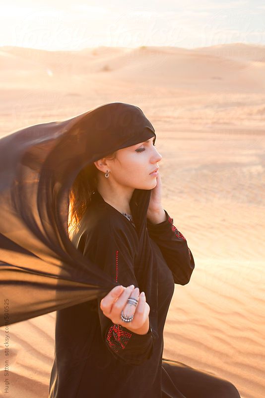 a woman in black is standing on the beach with her scarf over her head and looking off into the distance