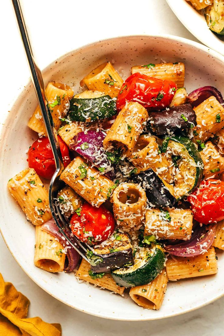 two bowls filled with pasta and vegetables on top of a white table next to bread