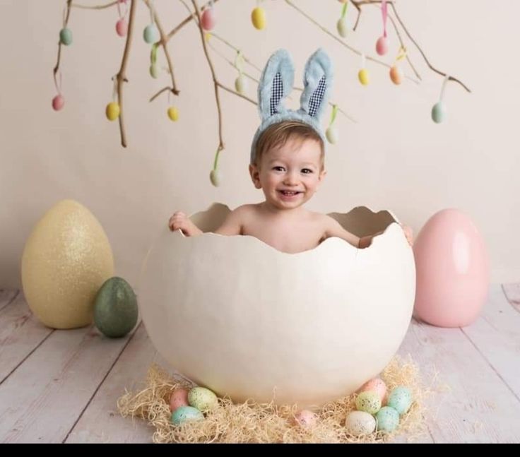 a baby sitting in an egg with bunny ears on his head, surrounded by easter decorations