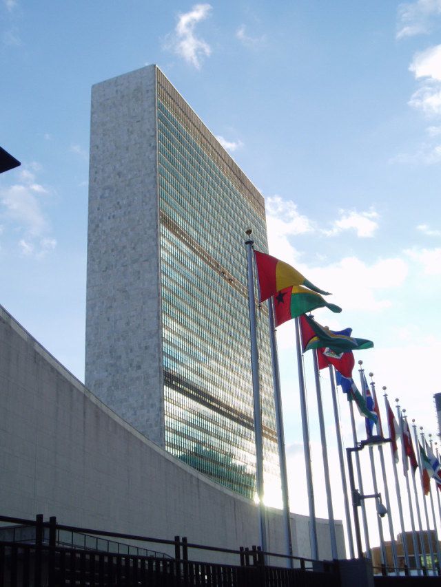 several flags are flying in front of a tall building with glass windows and a sky background