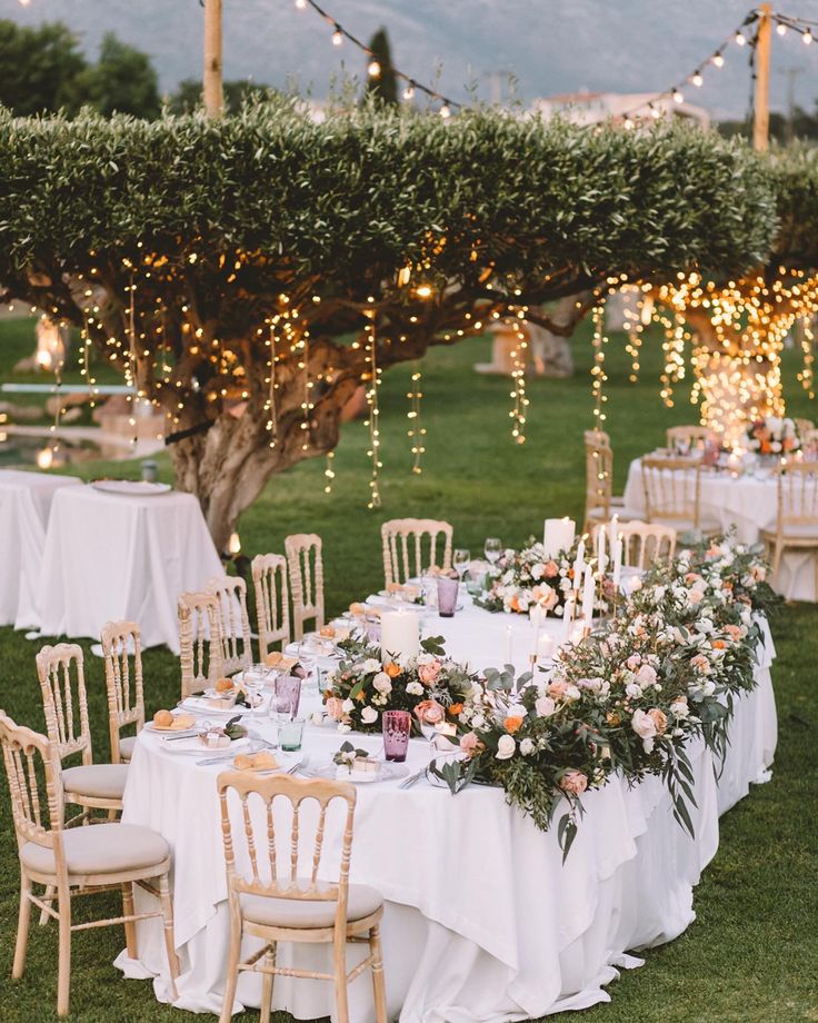 an outdoor dinner table set up with white linens and floral centerpieces, surrounded by string lights