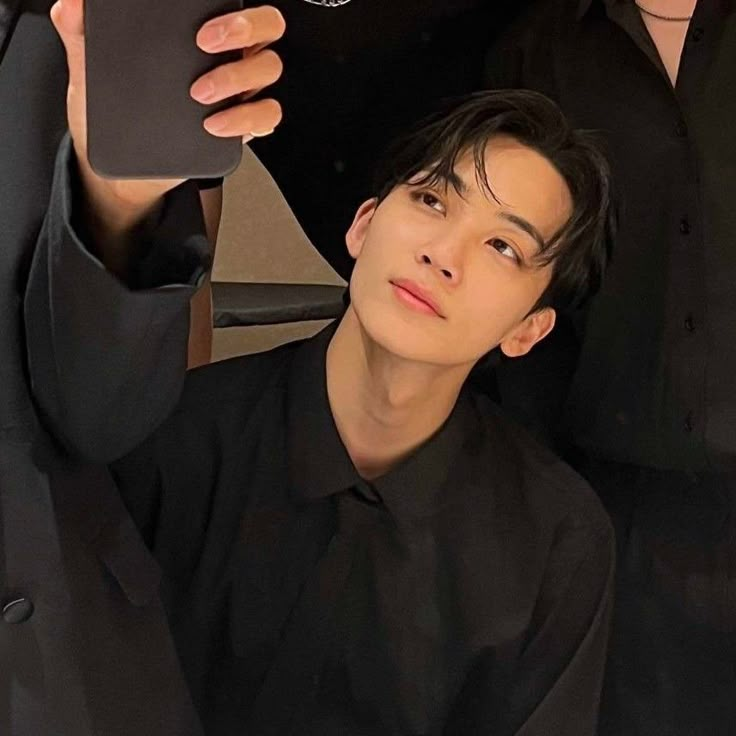 a young man holding up his cell phone to take a selfie with another person in the background