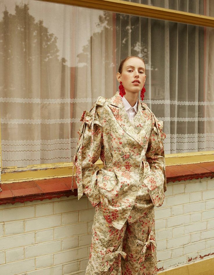 a woman standing in front of a window next to a brick wall wearing a suit and red earrings