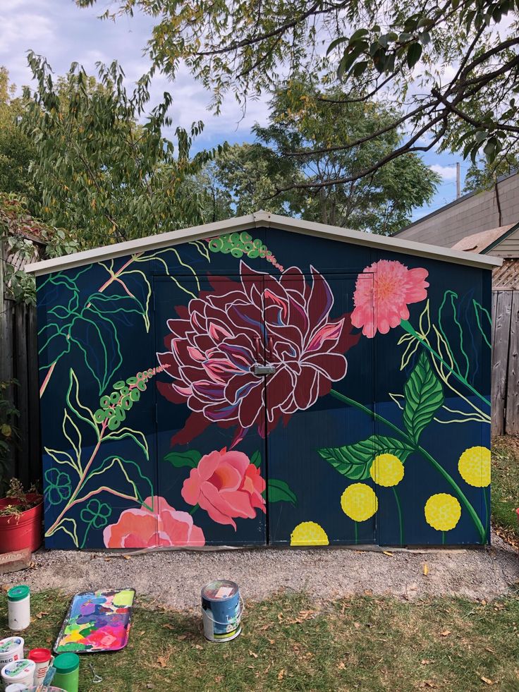 an outdoor painted shed with flowers on it and paint cans in the foreground next to it