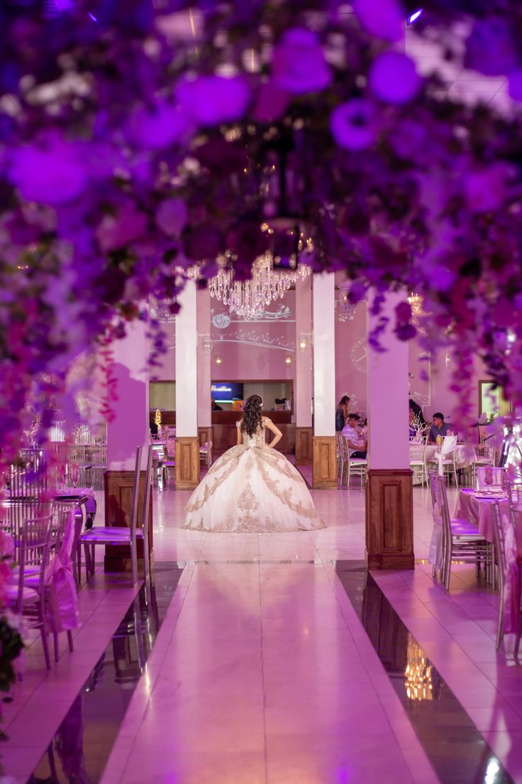 the bride is sitting in her wedding dress at the reception table with purple flowers hanging from the ceiling