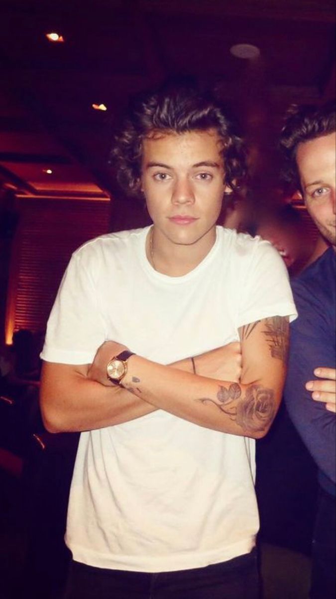 a young man with his arms crossed standing in a dark room, wearing a white t - shirt and black pants