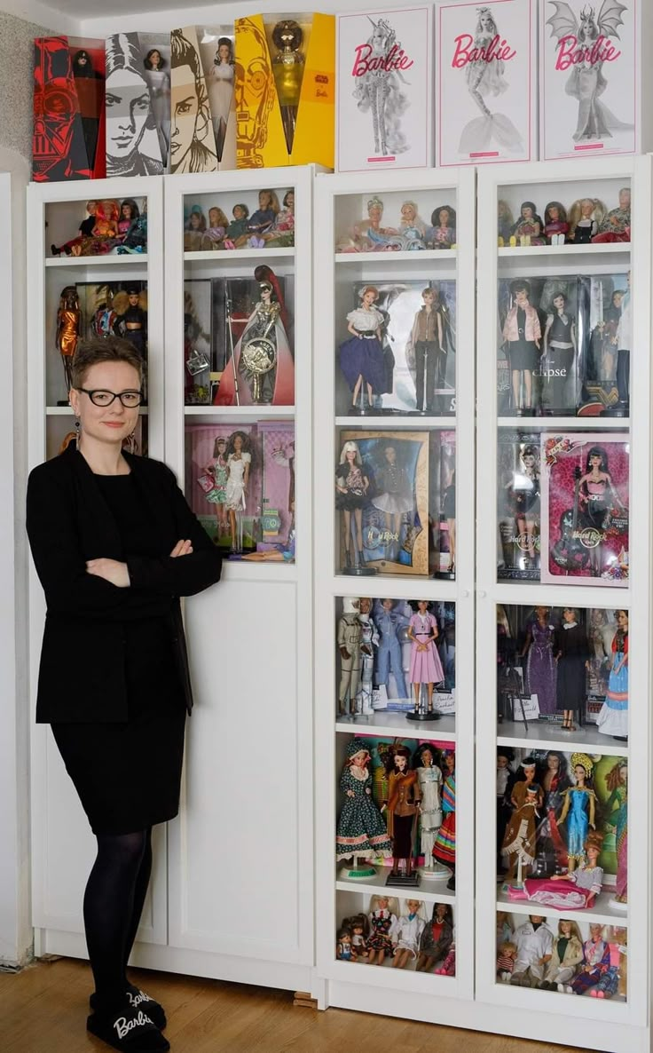 a woman standing in front of a bookcase filled with barbies and other dolls