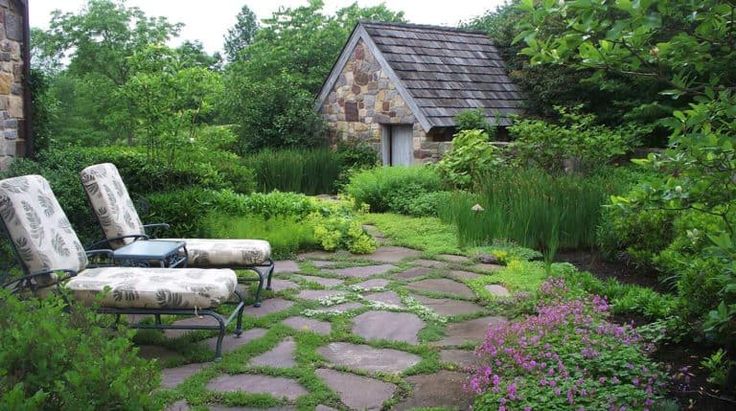 two lawn chairs sitting on top of a stone walkway next to flowers and trees in the background