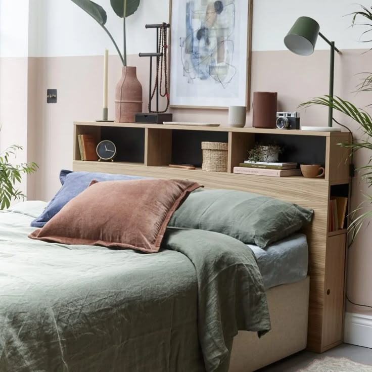 a bed with green sheets and pillows next to a book shelf filled with plants in front of a pink wall