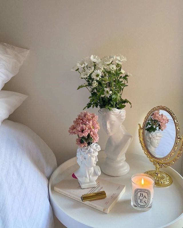 a white table with flowers and a mirror on it next to a candle, books and a pillow