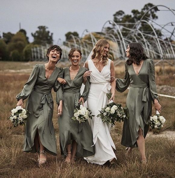 four bridesmaids in green dresses laughing and walking through an open field with their bouquets