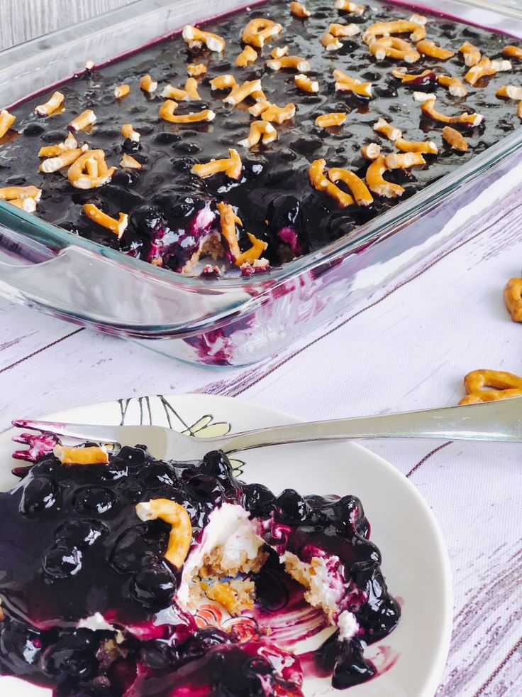 a blueberry dessert is served on a plate with a fork and glass casserole dish in the background