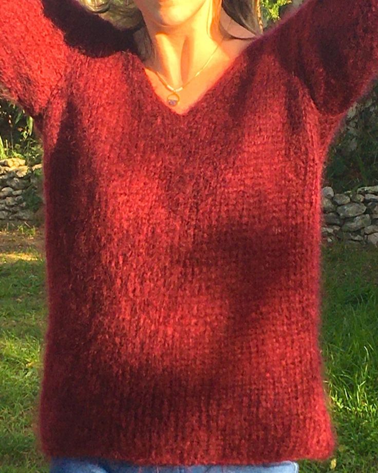 a woman standing in the grass with her arms behind her head, wearing a red sweater