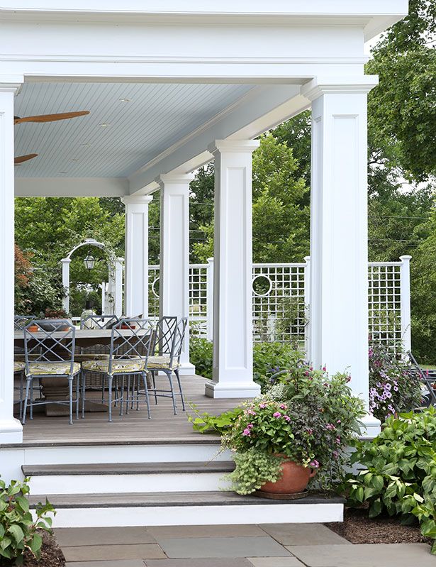 an outdoor dining area with white pillars and blue chairs on the front porch, surrounded by greenery