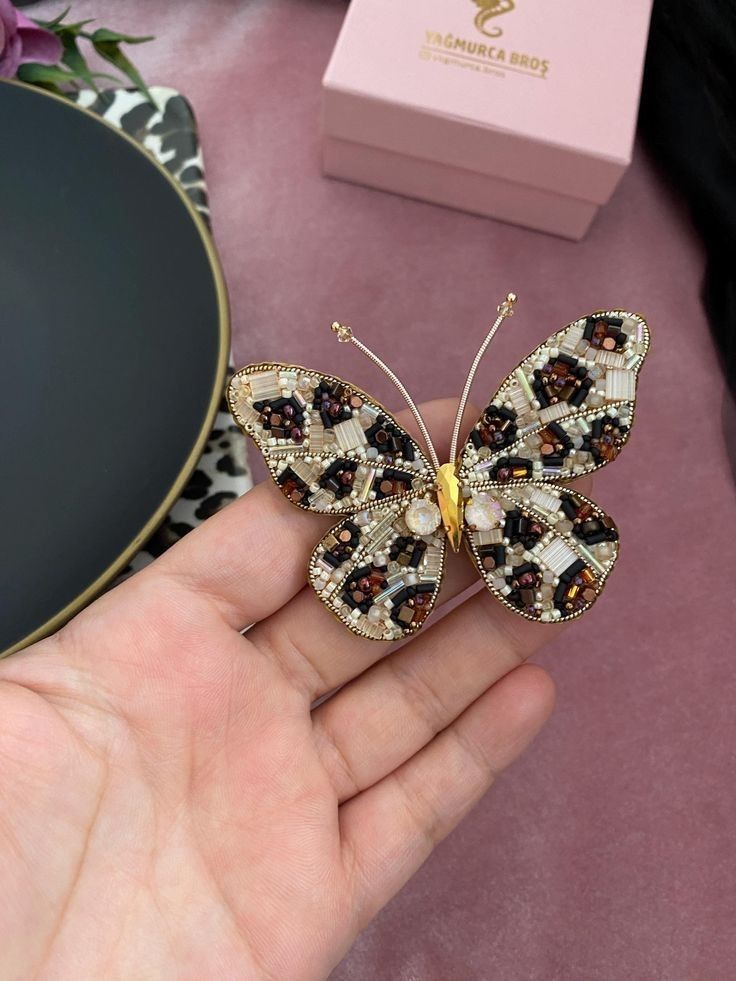 a hand holding a butterfly brooch in front of a pink box on a table