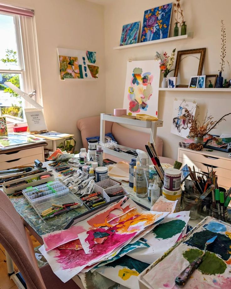 an artist's studio with lots of art supplies on the table and paintings on the wall