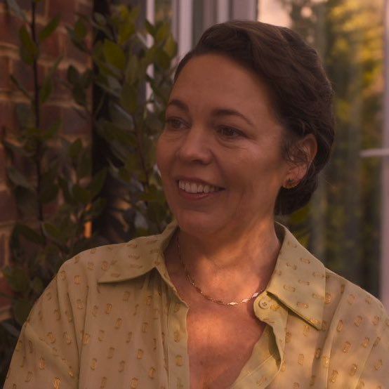 an older woman wearing a yellow shirt smiles at the camera while standing in front of a brick wall