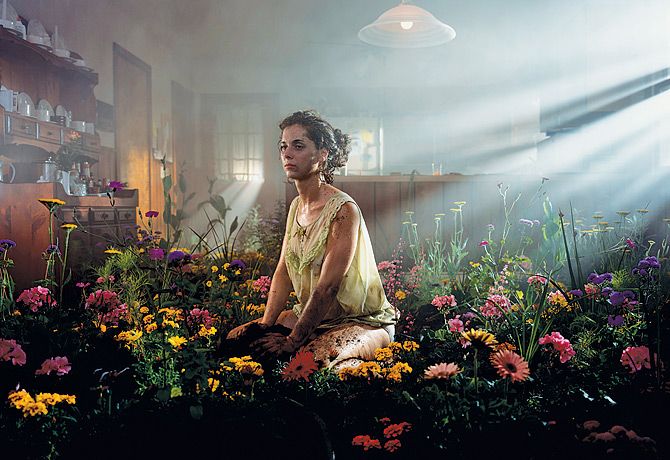 a woman sitting on the ground surrounded by flowers in front of a light that is coming from behind her