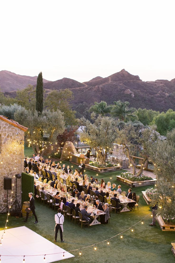 an outdoor event with people sitting at tables and lights on the grass, surrounded by mountains