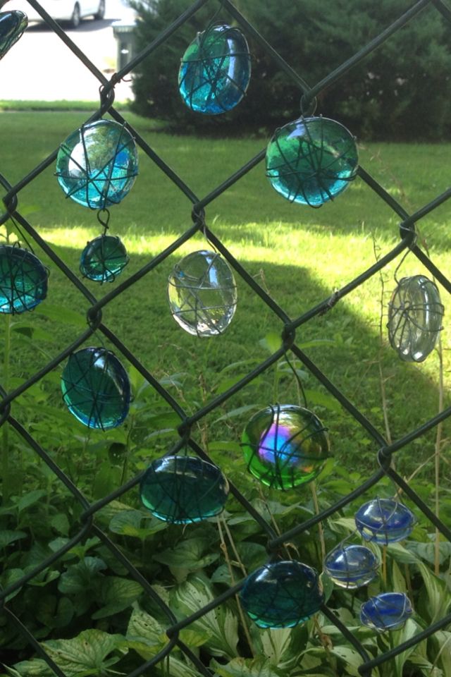 some glass balls are hanging on a chain link fence in the grass near trees and bushes