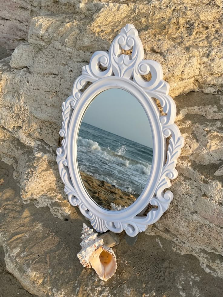 a mirror sitting on top of a rock next to the ocean with a shell in front of it