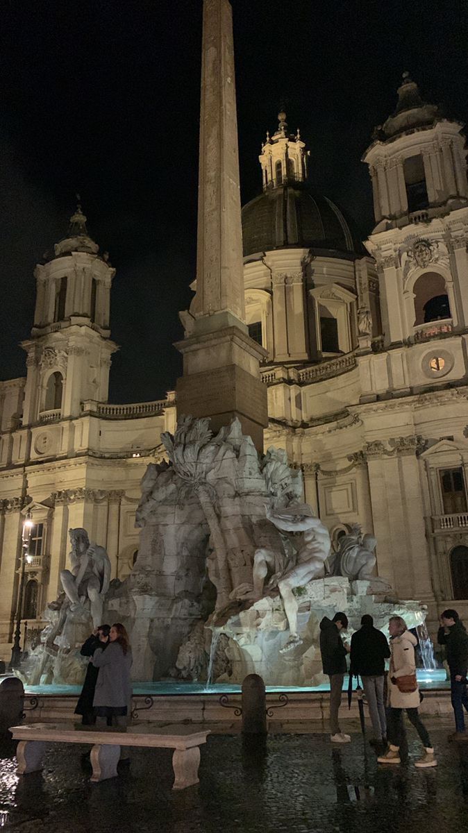 people are standing around in front of a building with a fountain and clock tower at night