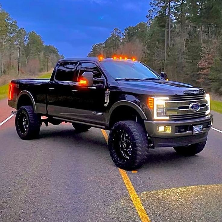 a large black truck with lights on driving down the road in front of some trees