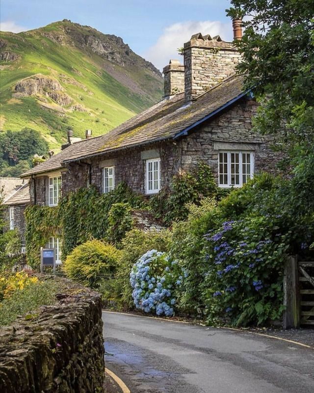 an old stone house surrounded by flowers and greenery with mountains in the back ground