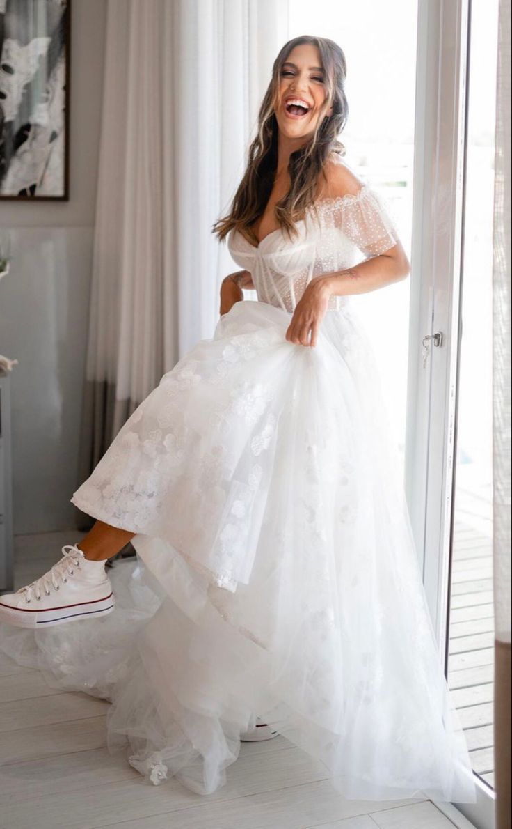 a woman in a white dress is standing by a window and smiling at the camera