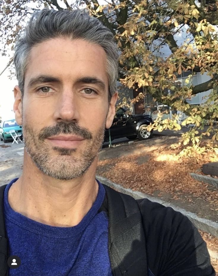 a man with grey hair and beard wearing a blue shirt is standing in front of a tree