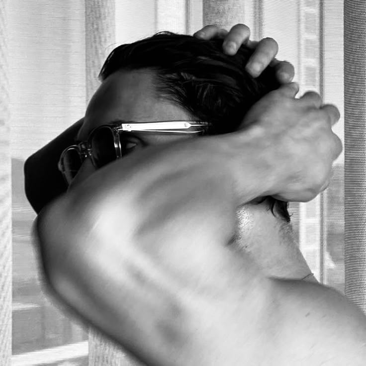 a man with no shirt is holding his hair in one hand and looking out the window