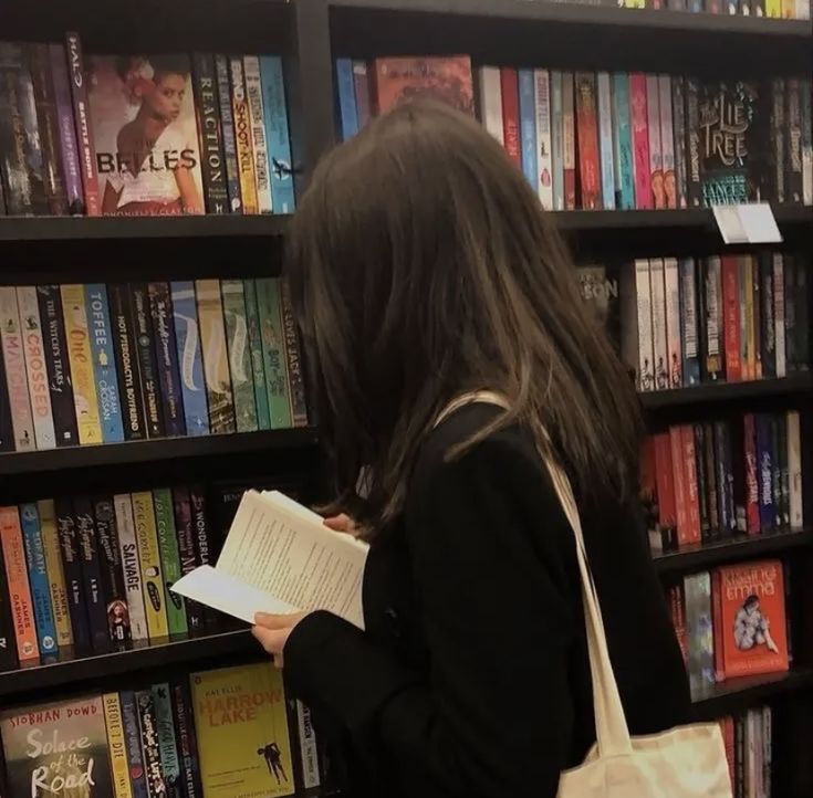 a woman standing in front of a bookshelf holding a book and looking at it