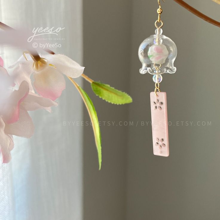 a pair of pink earrings hanging from a branch with flowers in the foreground and a window behind it