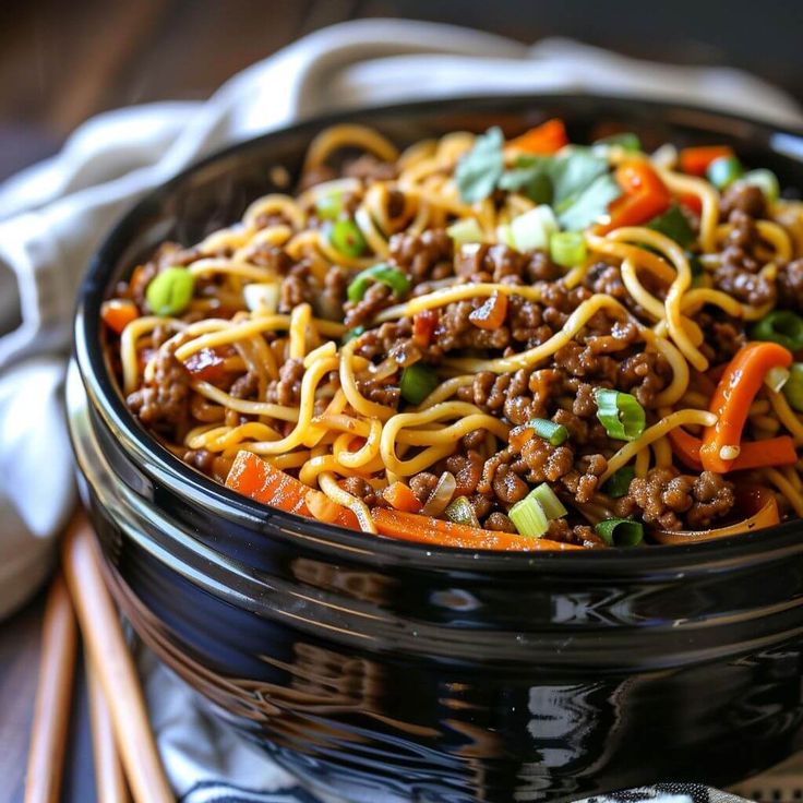 a black bowl filled with noodles and ground beef on top of a table next to chopsticks