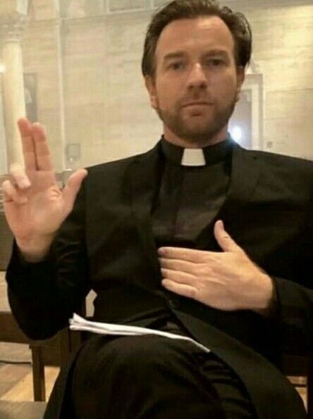 a man in a priest's outfit sitting down with his hand up to the side